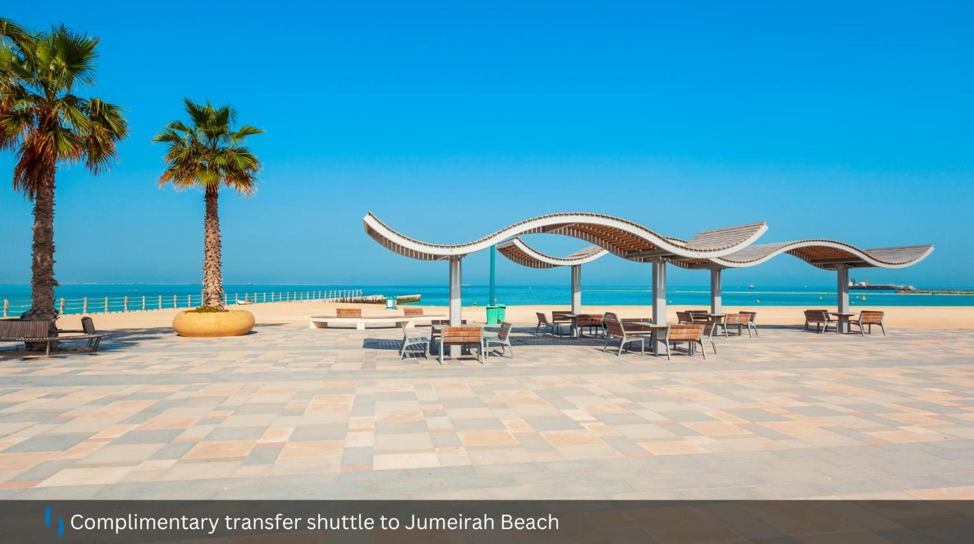 Grand Cosmopolitan Hotel Dubai Exterior foto The photo shows a scenic beach area, likely at Jumeirah Beach. In the foreground, there are wooden benches and tables arranged under shaded structures with a wavy design. A tall palm tree adds to the tropical feel of the setting. In the background, y